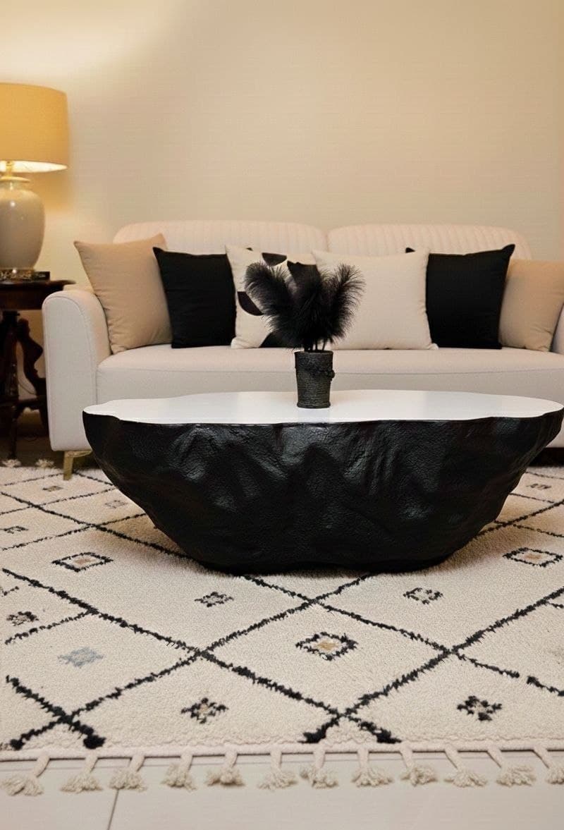 Sculptural organic coffee table featuring an irregular black textured craggy base and smooth flat off-white inset top, positioned on a patterned rug in front of an off-white sofa.