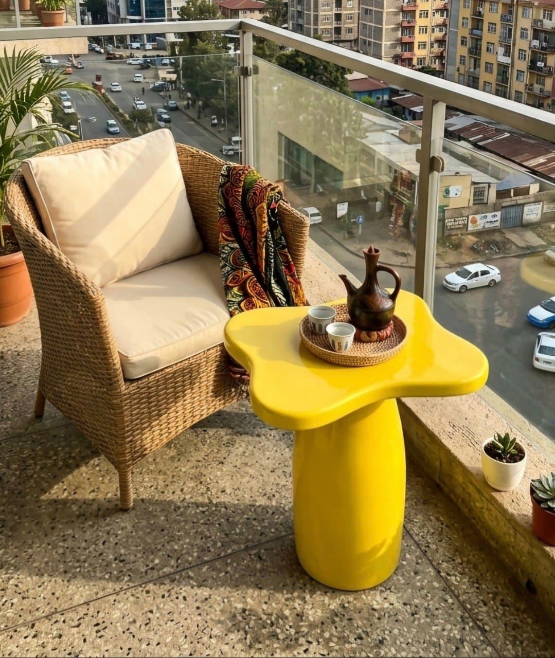Vibrant yellow clover-profile balcony side table in sun-baked ochre yellow gel coat holding traditional clay coffee pot and cups on a high-rise stone terrace next to a woven wicker chair with cream pillows